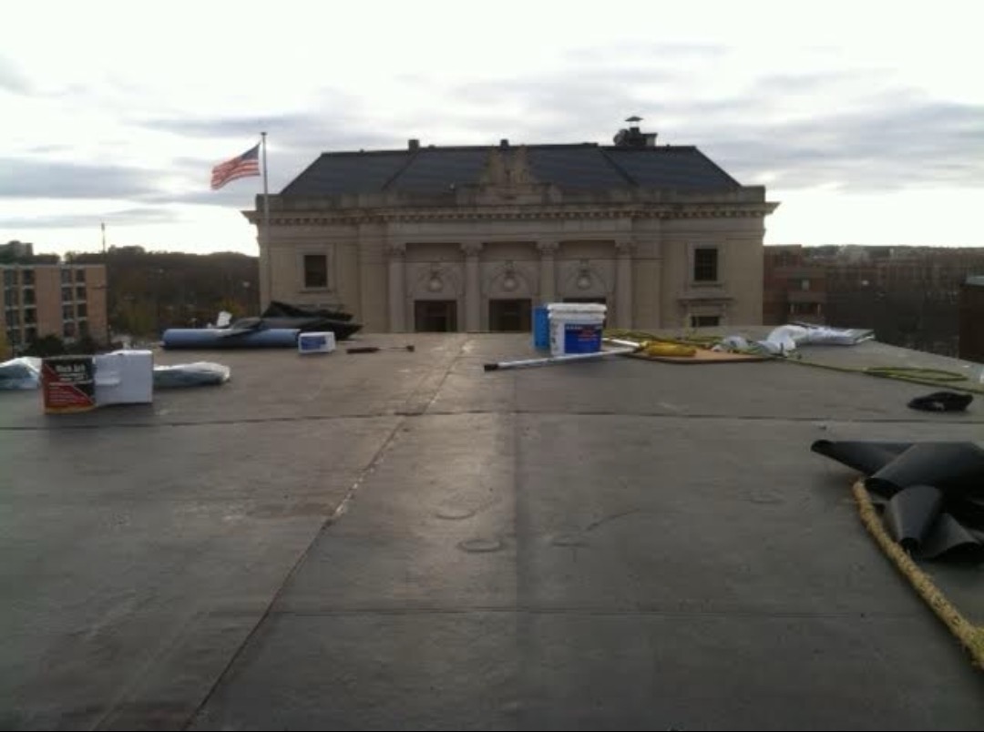 Flat commercial roof mid-install with rolls of rubber roofing, buckets of adhesive and tools laid out, historic stone building facade and American flag visible beyond — commercial and residential rubber roofing by Claflin Construction in Eau Claire