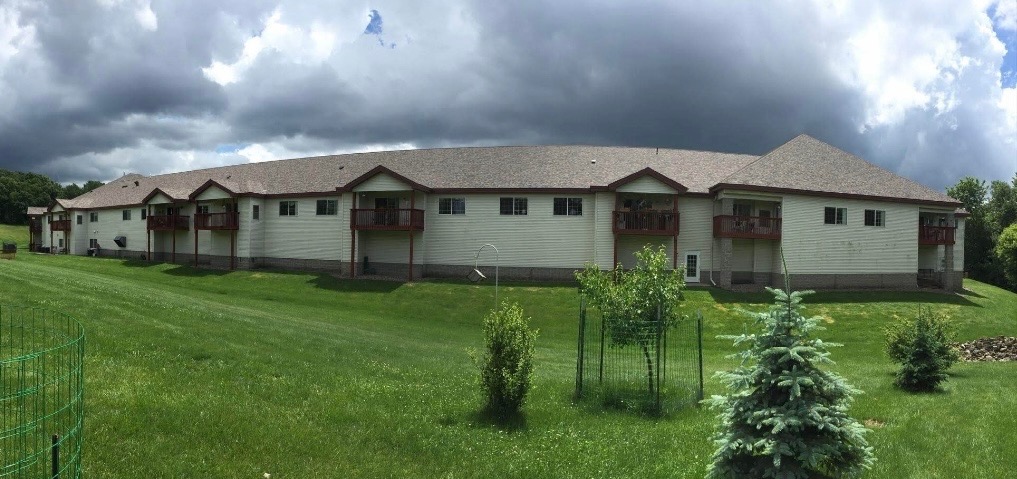 Large multi-unit residential building with new Owens Corning asphalt shingle roof stretching across a low-slope, hip-style roofline under dramatic storm clouds, green lawn and small trees in foreground indicating professional roofing replacement by Claflin Construction in Eau Claire