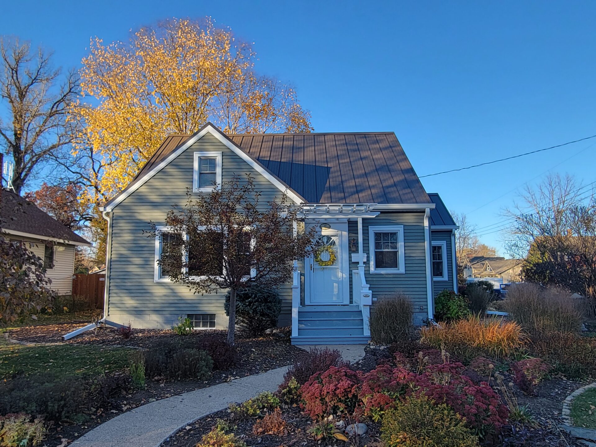 A blue-gray single-family home with new lap siding and a metal roof, centered front entrance with white trim and steps, fall landscaping and a winding walkway under a clear blue sky — siding and exterior upgrade for Eau Claire, Wisconsin.