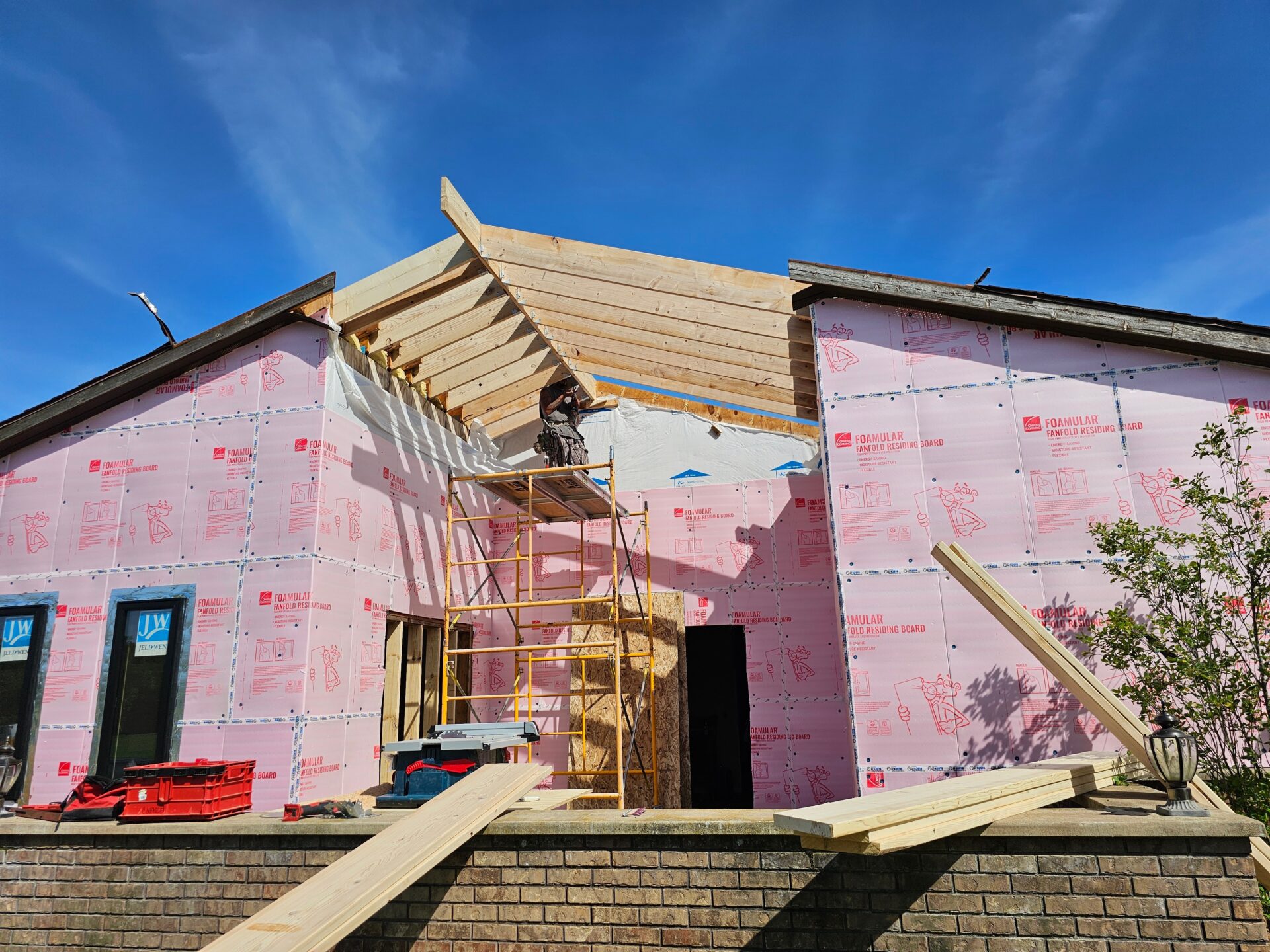 Residential construction site with pink foam sheathing installed on exterior walls, workers on scaffolding adding roof framing and insulation for improved energy efficiency and comfort
