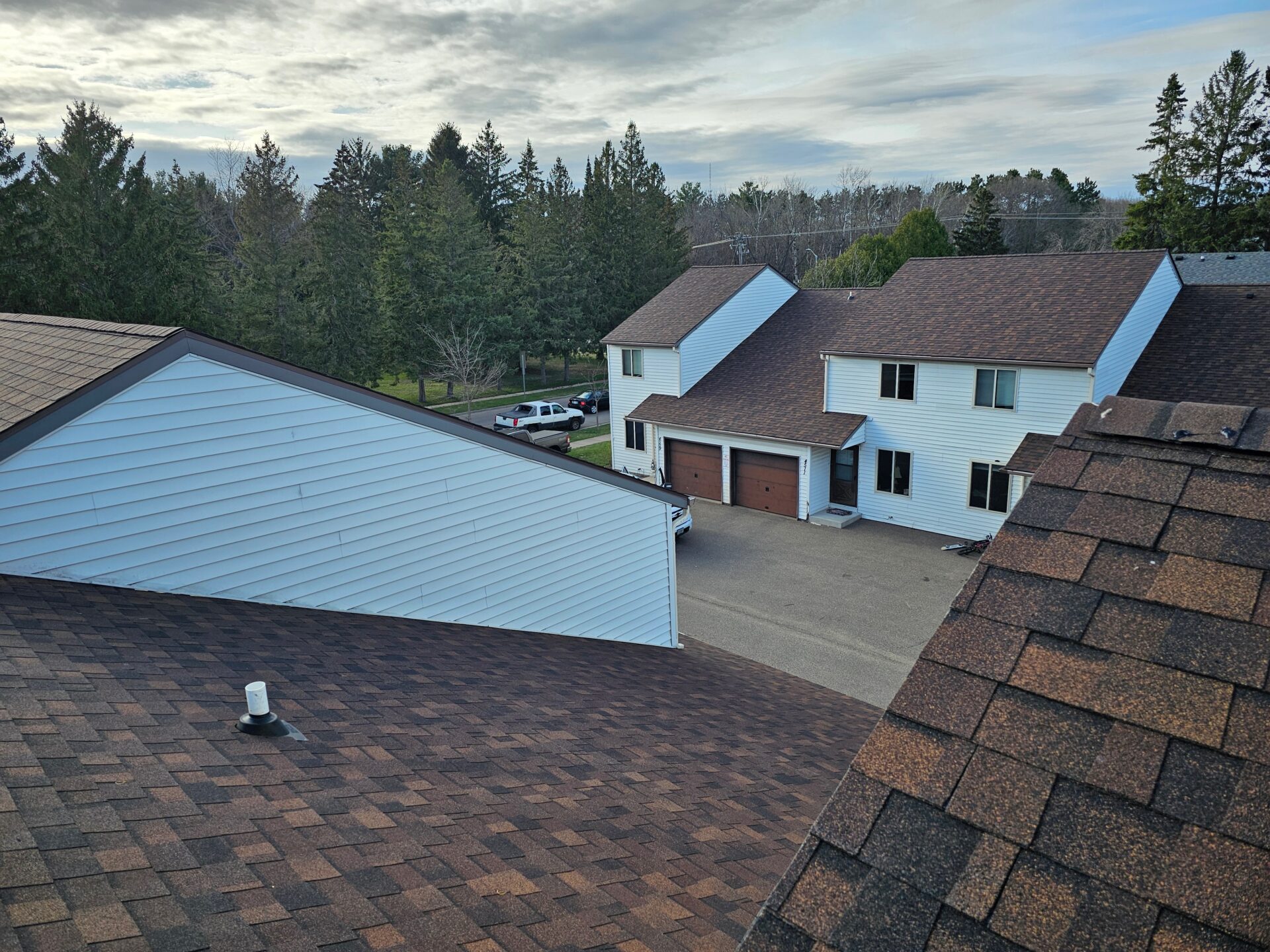 Residential neighborhood view from a rooftop showing newly installed brown Owens Corning asphalt shingle roofs on townhomes, adjacent garage buildings, roof vent flashing, and tree-lined background under a cloudy sky
