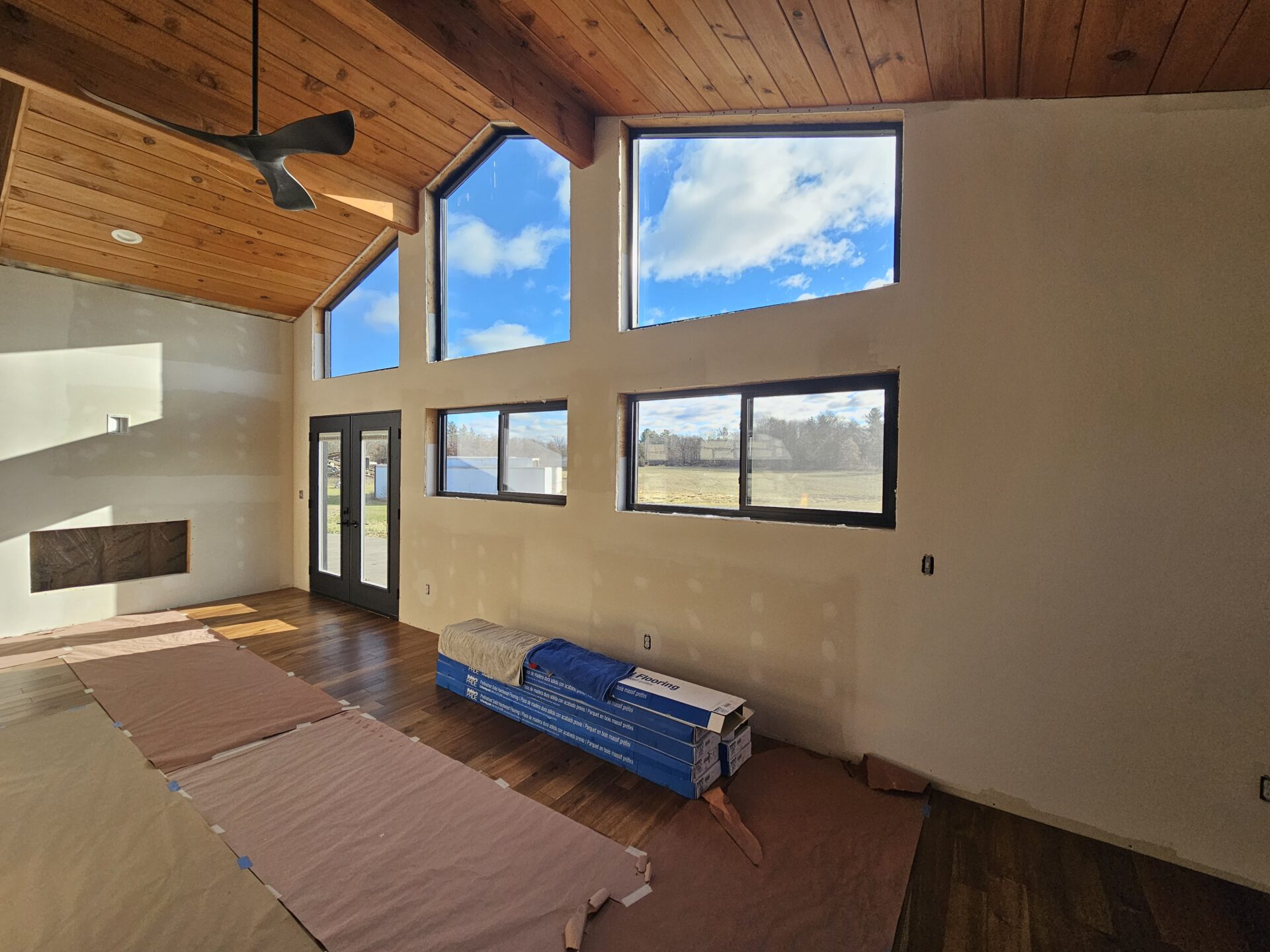 Sunlit living room under renovation with vaulted wood-paneled ceiling, modern black ceiling fan, tall triangular and rectangular windows showing blue sky, sliding glass doors, partially finished drywall, stacked flooring materials on protected hardwood floor — custom remodel by Eau Claire contractor.