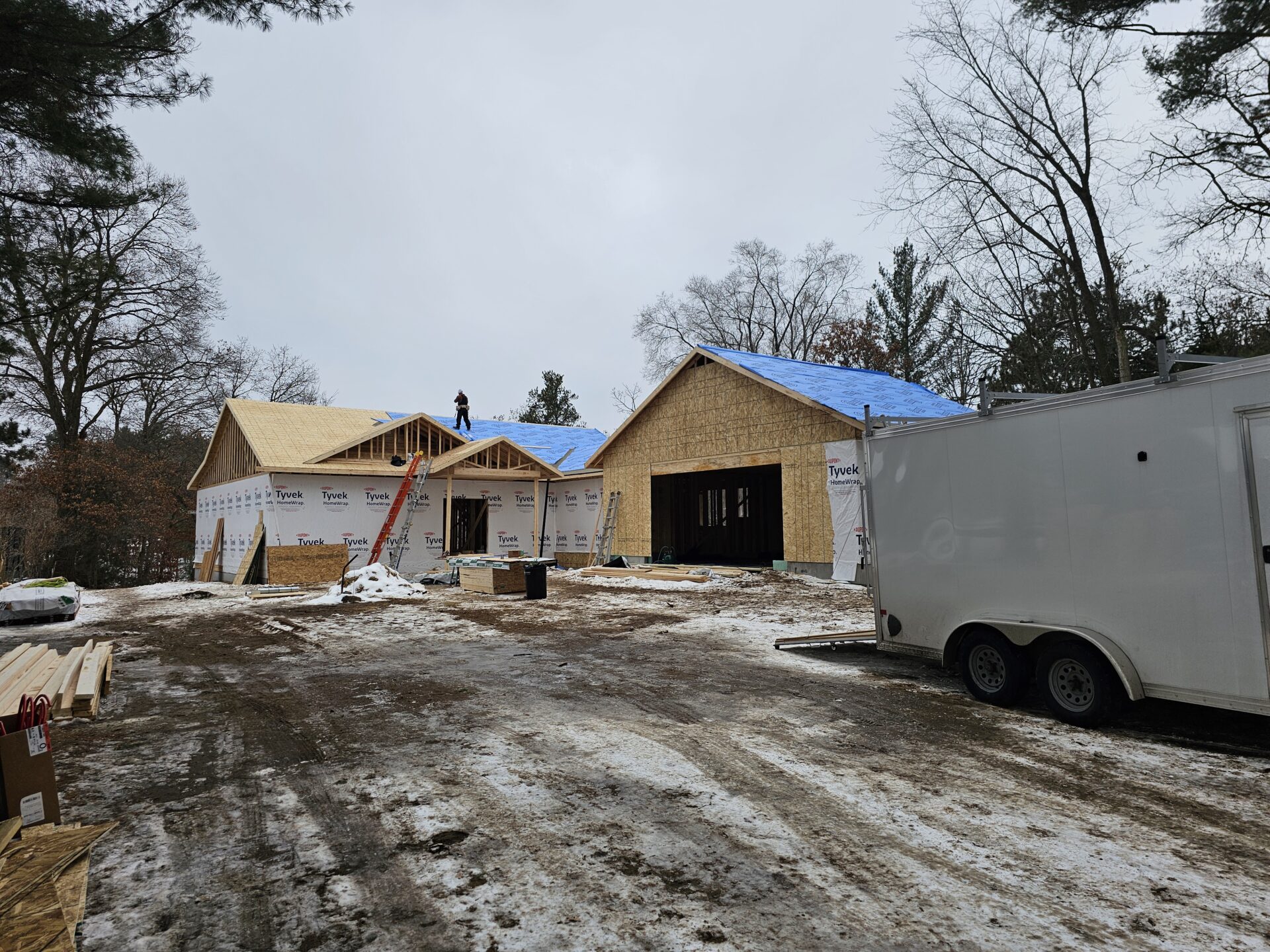 New home under construction with wood framing, Tyvek-wrapped exterior walls, blue roof underlayment and workers on the roof in a snowy Eau Claire jobsite — custom home construction and remodeling by Claflin Construction, licensed contractor