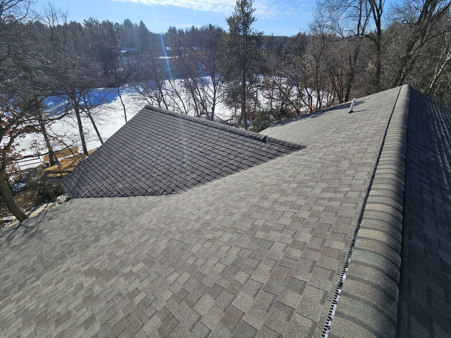 Aerial view of a newly shingled residential roof with ridge vent and cleanly sealed hips, overlooking a snowy tree-lined yard — professional roofing work by Claflin Construction in Eau Claire, WI