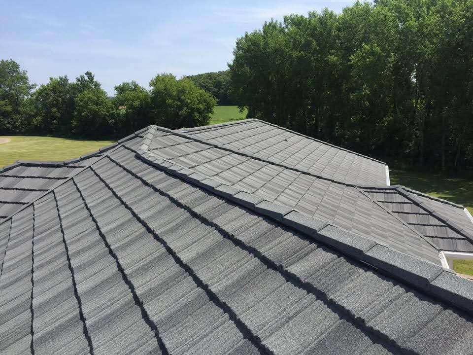 Close-up view of newly installed dark gray stone‑coated steel roofing panels on a residential multi‑ridge roof with surrounding green trees and lawn, showcasing durable, impact‑resistant roof system suitable for homeowners and commercial properties.